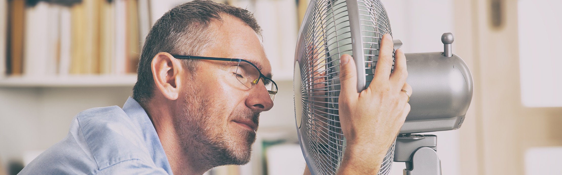Ein Mann mit Brille sitzt am Schreibtisch und hält sein Gesicht nah vor einen Ventilator zur Abkühlung  – Mehr erfahren zu Abkühlung im Sommer →